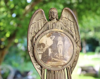 Op de afbeelding: Een vintage engelenbeeld met uitgestrekte vleugels, met een rond embleem met een religieuze scène. Het beeld heeft een bronzen afwerking en staat tegen een vage groene achtergrond. Het embleem heeft de tekst "N.D. LOURDES".