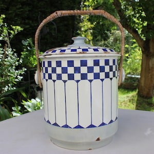 May include: A white enamelware pitcher with a blue and white chequered pattern and a woven handle. The pitcher has a lid and is sitting on a white surface.