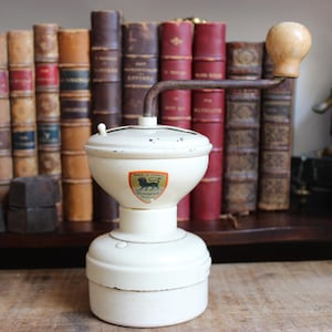 May include: Cream-colored vintage coffee grinder with a wooden handle and a shield emblem. The grinder is set against a backdrop of antique books with red and brown covers, creating a classic aesthetic.