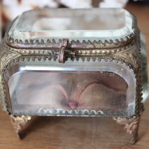 May include: A small, clear glass trinket box with a brass hinged lid and ornate feet. The box is empty and has a slightly aged appearance.