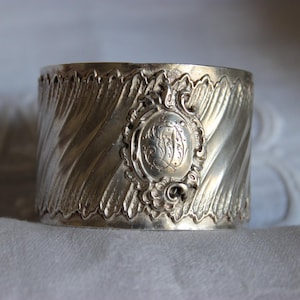 Pode incluir: Um porta-guardanapo prateado com um padrão detalhado em espiral. O anel apresenta uma cartela ovalada ornamentada com um monograma gravado. O design lembra a prataria antiga, adicionando um toque de elegância a qualquer mesa.