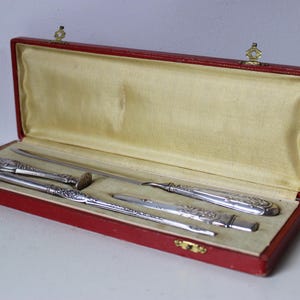 Op de afbeelding: Een rode leren doos met een gouden sluiting, met daarin een set van drie zilverkleurige gereedschappen. De gereedschappen zijn een briefopener, een zakmes en een klein schaartje.
