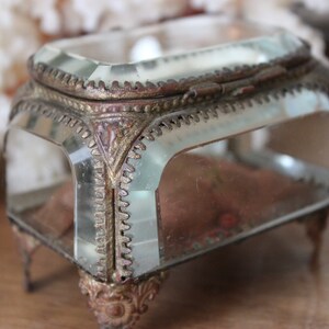 May include: A small, clear glass box with a gold metal frame and feet. The box has a hinged lid and is decorated with intricate details.