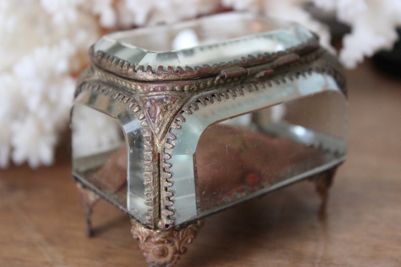 May include: A small, clear glass box with a gold metal frame and feet. The box has a hinged lid and is decorated with intricate details.