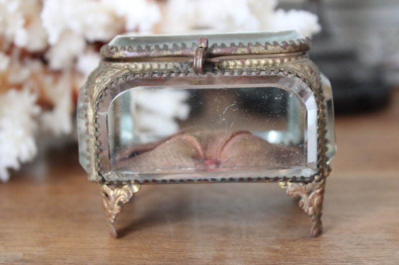 May include: A small, clear glass trinket box with a gold-toned metal frame and ornate feet. The box has a hinged lid and a velvet lining.
