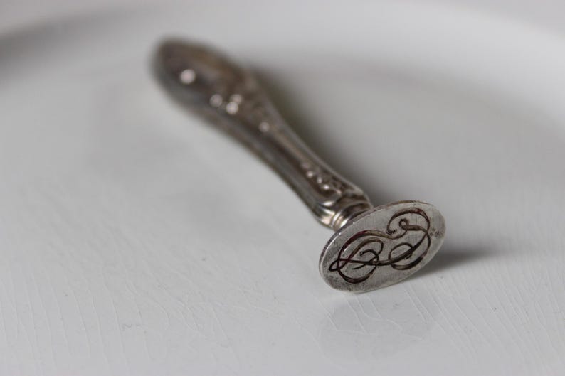 Op de afbeelding: Een zilveren zegel met een decoratief handvat en een ronde zegel met een monogram.