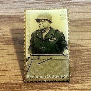 Può includere: Una spilla dorata a forma di francobollo, raffigurante Benjamin O. Davis, Sr. La spilla presenta la scritta "BLACK HERITAGE" a sinistra e "B2 USA" a destra. L'immagine mostra Davis in uniforme militare.