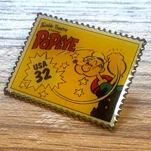 Op de afbeelding: Een gele Popeye-postzegel met een cartoonafbeelding van Popeye die zijn arm spant. De postzegel leest "Popeye" en "USA 32" met een gekartelde rand. De postzegel ligt op een houten oppervlak.
