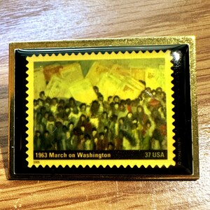 May include: A rectangular pin featuring a postage stamp design commemorating the 1963 March on Washington. The stamp has a yellow border and depicts a crowd of people. The pin has a gold-colored frame.
