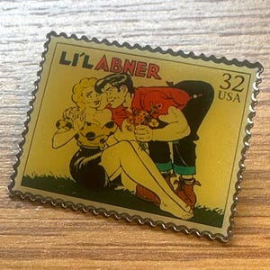 May include: A pin shaped like a postage stamp featuring a comic strip image of a man offering flowers to a woman. The stamp reads "LI'L ABNER" and "32 USA" in red text. The pin has a gold border and a yellow background.