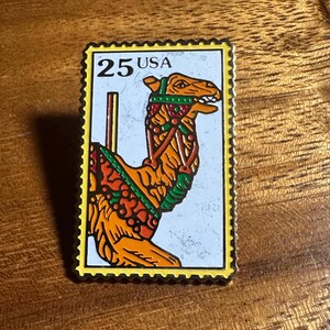 May include: A rectangular pin featuring a camel design, resembling a postage stamp. The pin has a yellow border with the text "25 USA". The camel is orange with green and red accents, set against a light blue background.