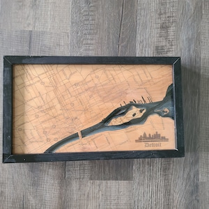 May include: A wooden map of Detroit, Michigan, with a black frame. The map is raised and shows the city's streets and waterways. The city skyline is depicted in the lower right corner, with the word "Detroit" below it.