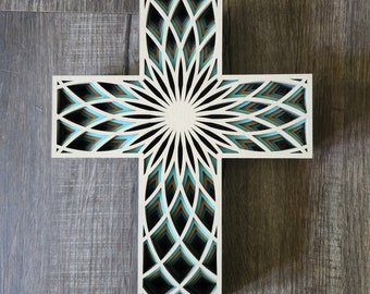 May include: A layered wooden cross with a detailed, geometric design. The cross features a cream-colored outer frame with intricate cutouts revealing layers of teal, brown, and black. The center has a sunburst pattern.