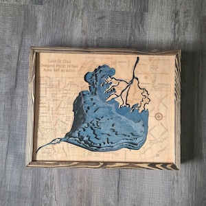 May include: A wooden map of Lake St. Clair with a blue inlay showing the lake's shape. The map is framed in a dark wood frame. The text on the map reads "Lake St. Clair, Deepest Point: 19 feet, Area: 440 sq miles."