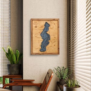 May include: A wooden wall art piece depicting a lake map with blue water and brown land features. The map is framed in a natural wood frame and is hanging on a beige wall.