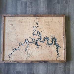 May include: Wooden framed map of Table Rock Lake in Missouri, Arkansas. The lake is depicted in blue against a light wood background. The map includes text with lake details and is framed in a dark brown wood.