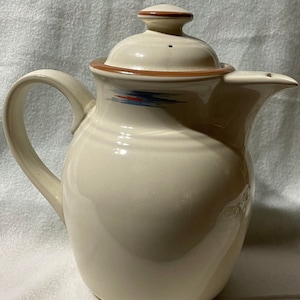 May include: A cream-colored ceramic coffee pot with a brown lid and a blue and red design. The pot has a rounded body and a handle on the left side.