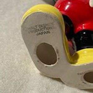 May include: A ceramic figurine of Mickey Mouse, standing with his arms outstretched. He is wearing his signature red shorts with white polka dots, yellow shoes, and black gloves. The figurine is made in Japan and has a hole in the bottom of his foot.