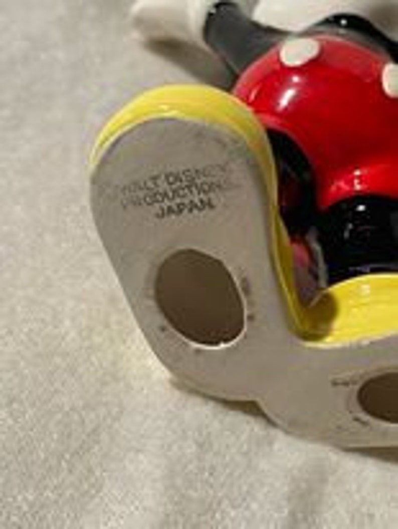May include: A ceramic figurine of Mickey Mouse, standing with his arms outstretched. He is wearing his signature red shorts with white polka dots, yellow shoes, and black gloves. The figurine is made in Japan and has a hole in the bottom of his foot.