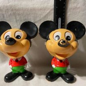 May include: Two vintage plastic Mickey Mouse figurines. Both are wearing red shirts with green pants and have black shoes. The figurines are approximately 5 inches tall.