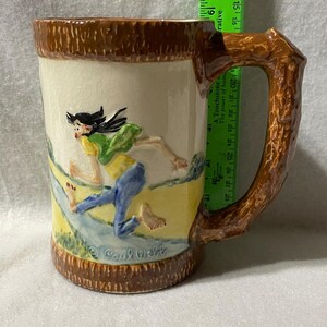 May include: Ceramic mug with a brown textured handle and rim. The mug features a relief design of a person running through a stream, with the text "Paul Webb" at the bottom. The mug is approximately 5 inches tall.