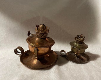 May include: Two antique oil lamps. The larger lamp is copper-toned with a handle and a circular base. The smaller lamp is brass-toned, also with a handle and a circular base. Both lamps have a crown-shaped wick holder.