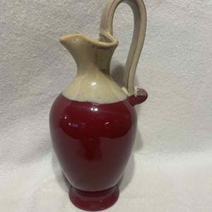 May include: A ceramic pitcher with a curved handle. The pitcher has a two-tone design, with a deep red base and a cream-colored top. The handle is cream-colored with red accents. The pitcher has a small spout.