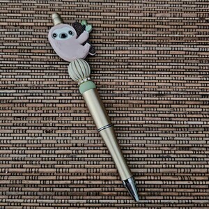 May include: A gold pen with a pink sloth charm and green and white beads. The pen is on a brown woven surface.