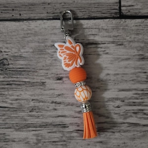 May include: Orange and white butterfly keychain with a silver clasp and a tassel. The butterfly has a white outline and orange wings. The keychain has a white and orange patterned bead and a silver bead.
