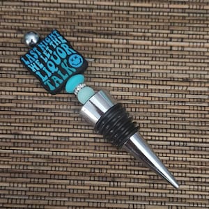 May include: A silver bottle stopper with a black rubber stopper and a blue and black charm with the text "Last night we let the liquor talk".