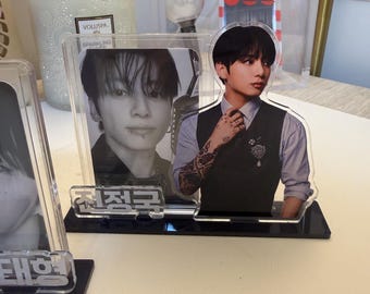 May include: A clear acrylic standee featuring a portrait of a person with dark hair, wearing a light shirt and dark vest, with visible arm tattoos. The base is black and displays the Korean text 전정국. A second, smaller black and white photo is attached.