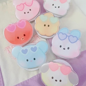 May include: A collection of seven acrylic phone grips featuring cartoon animal faces in pastel colors. Each character wears heart-shaped glasses in various shades including pink, blue, and green. The designs include bears, rabbits, and dogs.