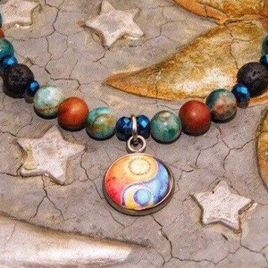 May include: A beaded necklace featuring a pendant with a yin-yang design. The necklace has a mix of colorful beads, including red, turquoise, and black, with blue accents. The pendant is round with a colorful design. The necklace is displayed on a textured surface.