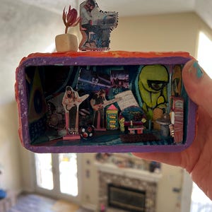 May include: A miniature room made of cardboard and painted with bright colors. The room is decorated with cut-out images of people playing music, a green alien, and a potted plant. The room is held in a hand.