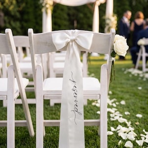 May include: A white wooden folding chair featuring a long, smooth white satin ribbon tied in a bow at the top. The ribbon hangs down the centre of the backrest and displays the word reserved written in a black, flowing cursive script font.