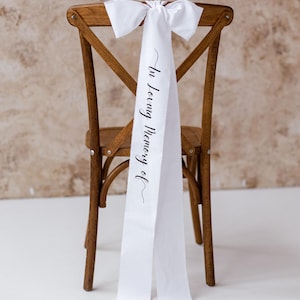May include: A wooden chair with a white satin sash tied to the back. The sash reads "In Loving Memory of" in black script. The chair is brown and has a cross-back design.