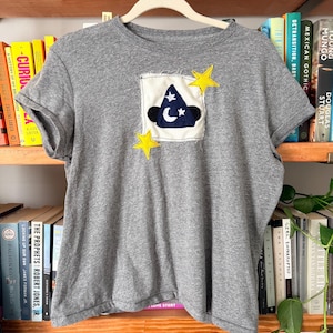 May include: A gray t-shirt with a patch featuring a wizard hat, moon, and stars. The patch is on a white square with a frayed edge, and yellow star accents are sewn on the shirt. The shirt is hanging on a white hanger.