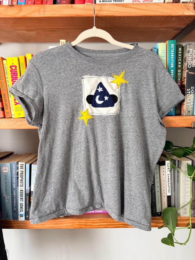 May include: A gray t-shirt with a patch featuring a wizard hat, moon, and stars. The patch is on a white square with a frayed edge, and yellow star accents are sewn on the shirt. The shirt is hanging on a white hanger.