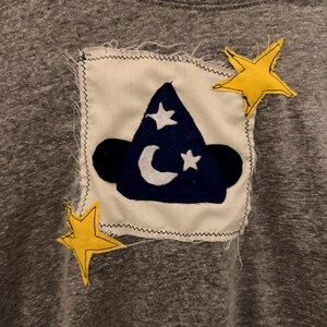 May include: Gray t-shirt with a sewn-on patch featuring a dark blue wizard hat with a white crescent moon and stars. Two yellow stars are sewn on the shirt near the patch. The patch is on a white background.