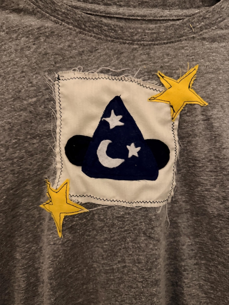May include: Gray t-shirt with a sewn-on patch featuring a dark blue wizard hat with a white crescent moon and stars. Two yellow stars are sewn on the shirt near the patch. The patch is on a white background.