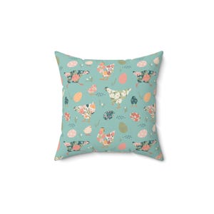 Op de afbeelding: Vierkant kussen met een herhalend patroon van kippen, kuikens en paaseieren op een turquoise achtergrond. De kippen en eieren zijn versierd met bloemmotieven in roze, oranje en groen.