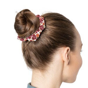 Può includere: Un elastico per capelli con un motivo di melograni rossi e arancioni su sfondo bianco. L'elastico è avvolto attorno a uno chignon di capelli castani. L'immagine è una vista laterale della testa e della spalla di una persona.