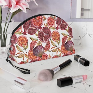 Può includere: Pochette per cosmetici con sfondo bianco e motivo di melograni, fichi e fette d'arancia. La borsa ha una cerniera nera e un cinturino da polso nero. Pennelli per il trucco e rossetti sono davanti alla borsa.