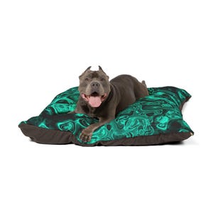 May include: A gray dog rests on a dog bed. The bed has a dark brown base and a top with a swirling teal and black pattern. The dog is looking at the camera with its tongue out.