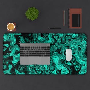 May include: An overhead view of a desk setup featuring a large desk mat with a teal and black swirling pattern. A laptop, mouse, coffee cup, notebook, pen, and small plant are arranged on the desk.