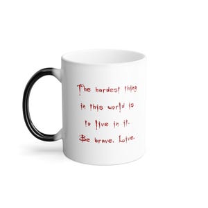 Może przedstawiać: Biały ceramiczny kubek z czarnym uchwytem, który przechodzi w biały. Kubek ma czerwony tekst: "The hardest thing in this world is to live in it. Be brave. Live."