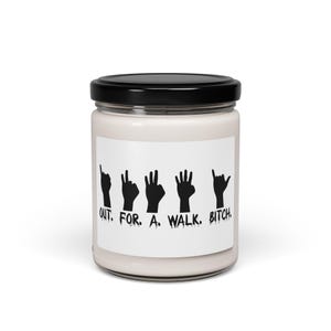 May include: A white candle jar with a black lid. The label features black hand gestures spelling out "OUT. FOR. A. WALK. BITCH."