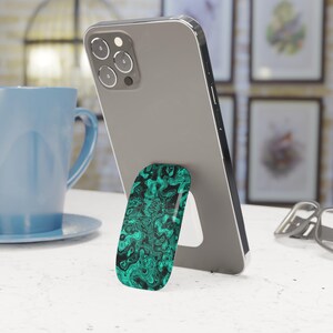 May include: A gray smartphone featuring a collapsible phone grip attached to the back. The grip has an oval shape and displays a vibrant, swirling pattern of black and bright teal colors that resemble liquid marble or abstract flowing textures.