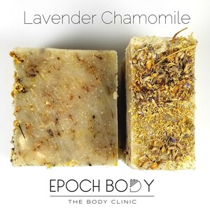Op de afbeelding: Twee stukken zeep, een met een lichtbruine kleur en de andere met een lichtgele kleur. Beide stukken zeep hebben gedroogde bloemen en kruiden erin verwerkt. De stukken zeep zijn gelabeld met "Lavender Chamomile" en "Epoch Body, The Body Clinic".