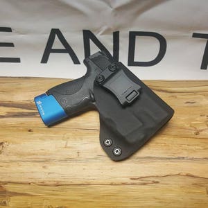 May include: A black leather holster with a blue gun inside. The holster has a clip on the back and is designed to be worn on a belt.